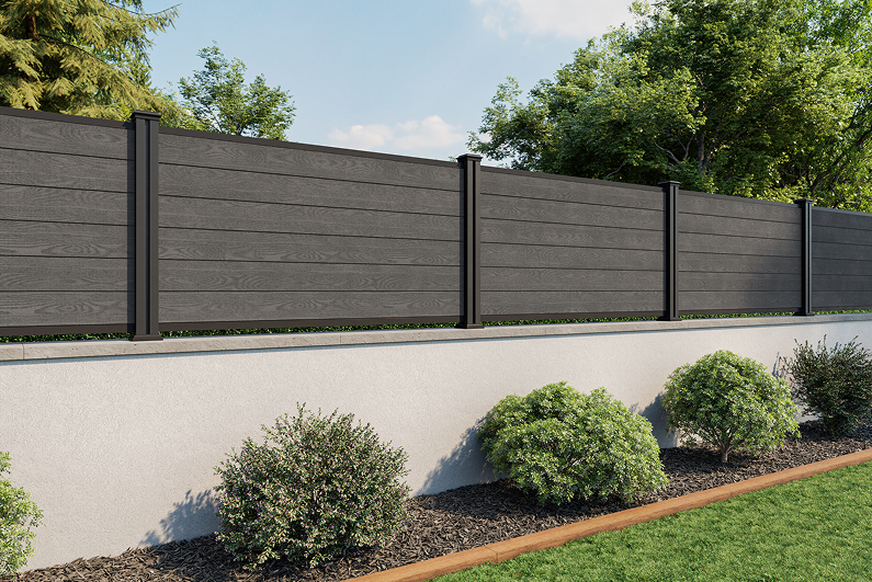 Modern dark grey composite fence on a light concrete wall with shrubs in front.